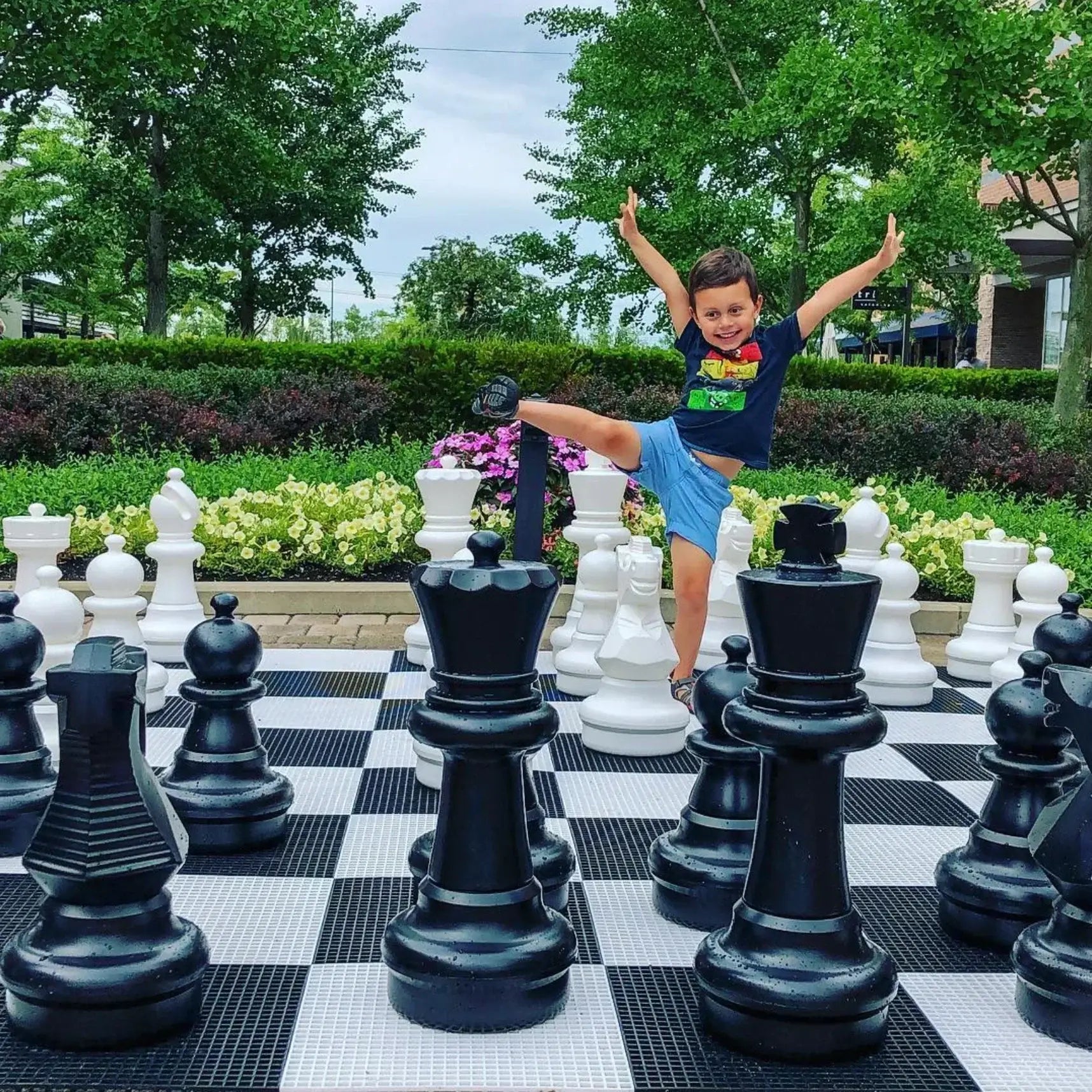 young-boy-jumping-with-arms-up-on-a-giant-chess-board-surrounded-by-black-and-white-pieces-showing-that-chess-is-fun-in-a-park-setting