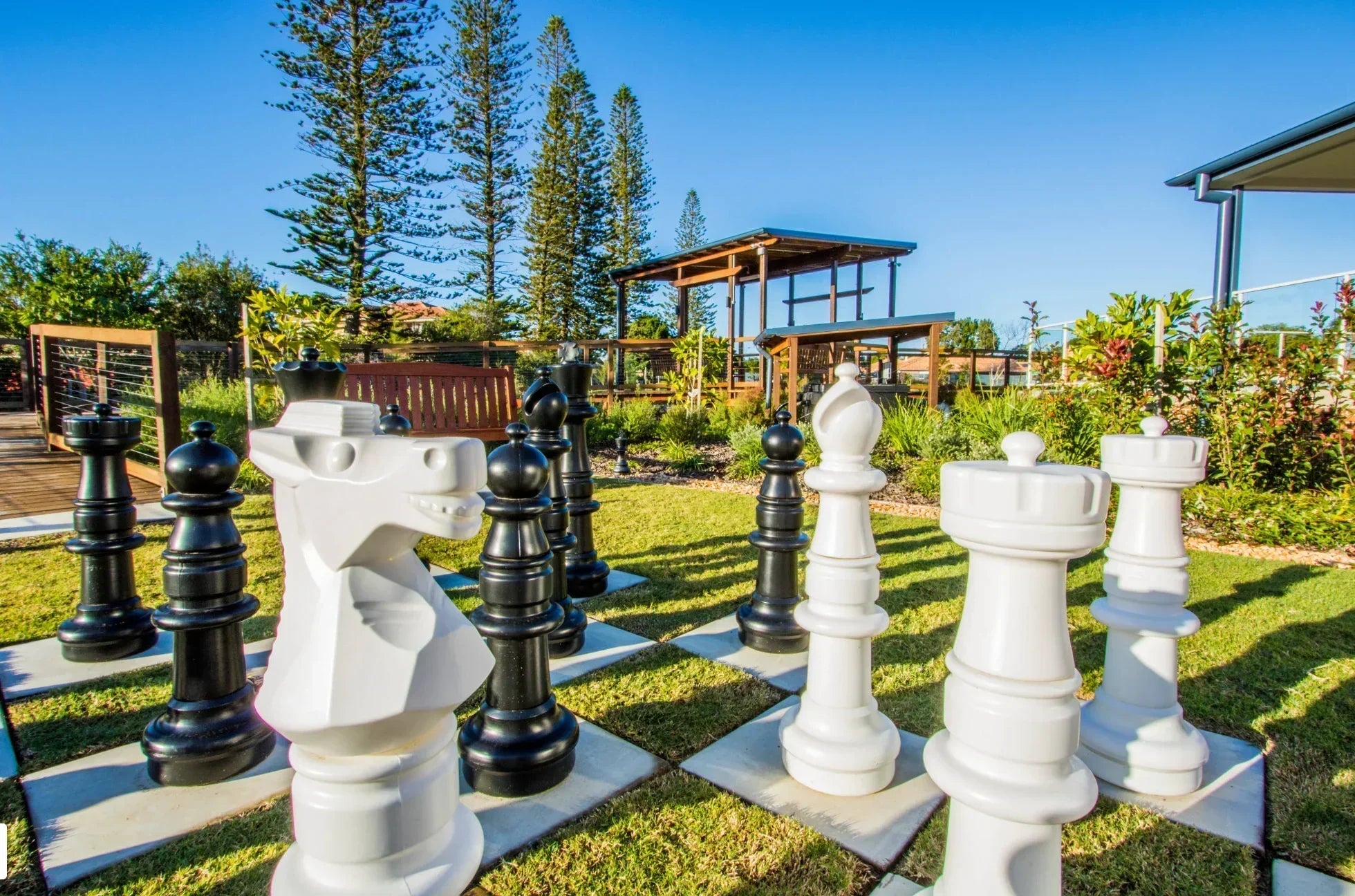 giant-black-and-white-chess-pieces-arranged-on-outdoor-grass-board-with-trees-wooden-bench-and-garden-structures-in-background-under-clear-blue-sky