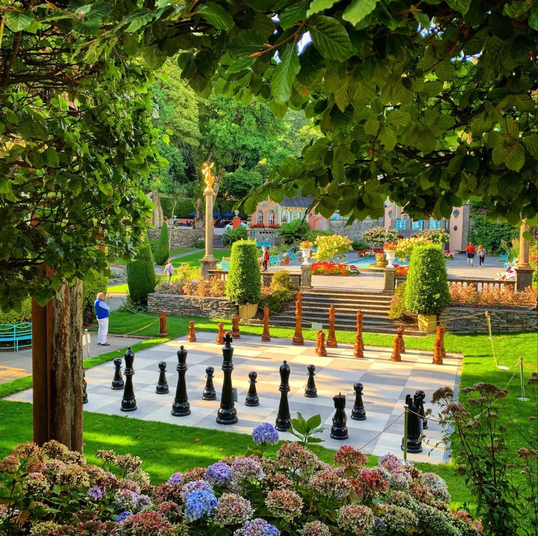 Giant outdoor chessboard with black and brown pieces in beautiful garden surrounded by flowers trees and decorative architecture on a sunny day.