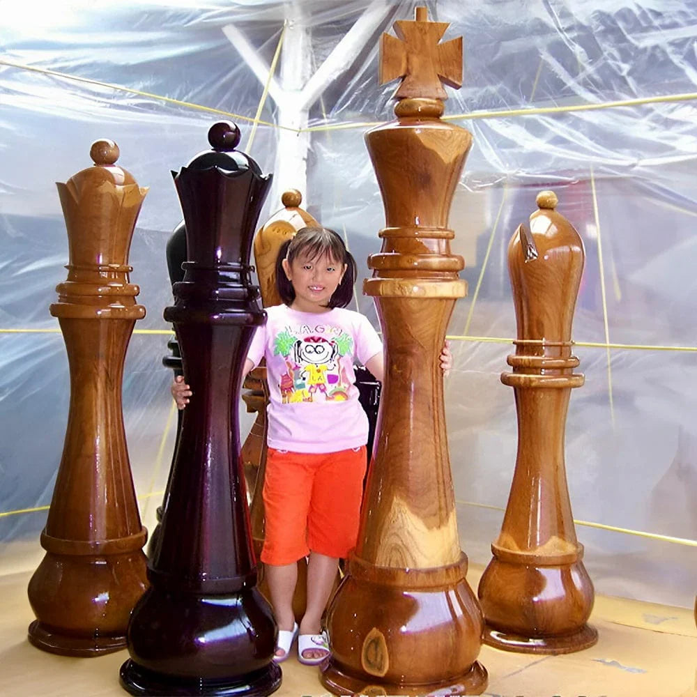 Young girl standing between giant wooden chess pieces including king queen and bishops with clear protective sheeting in background.