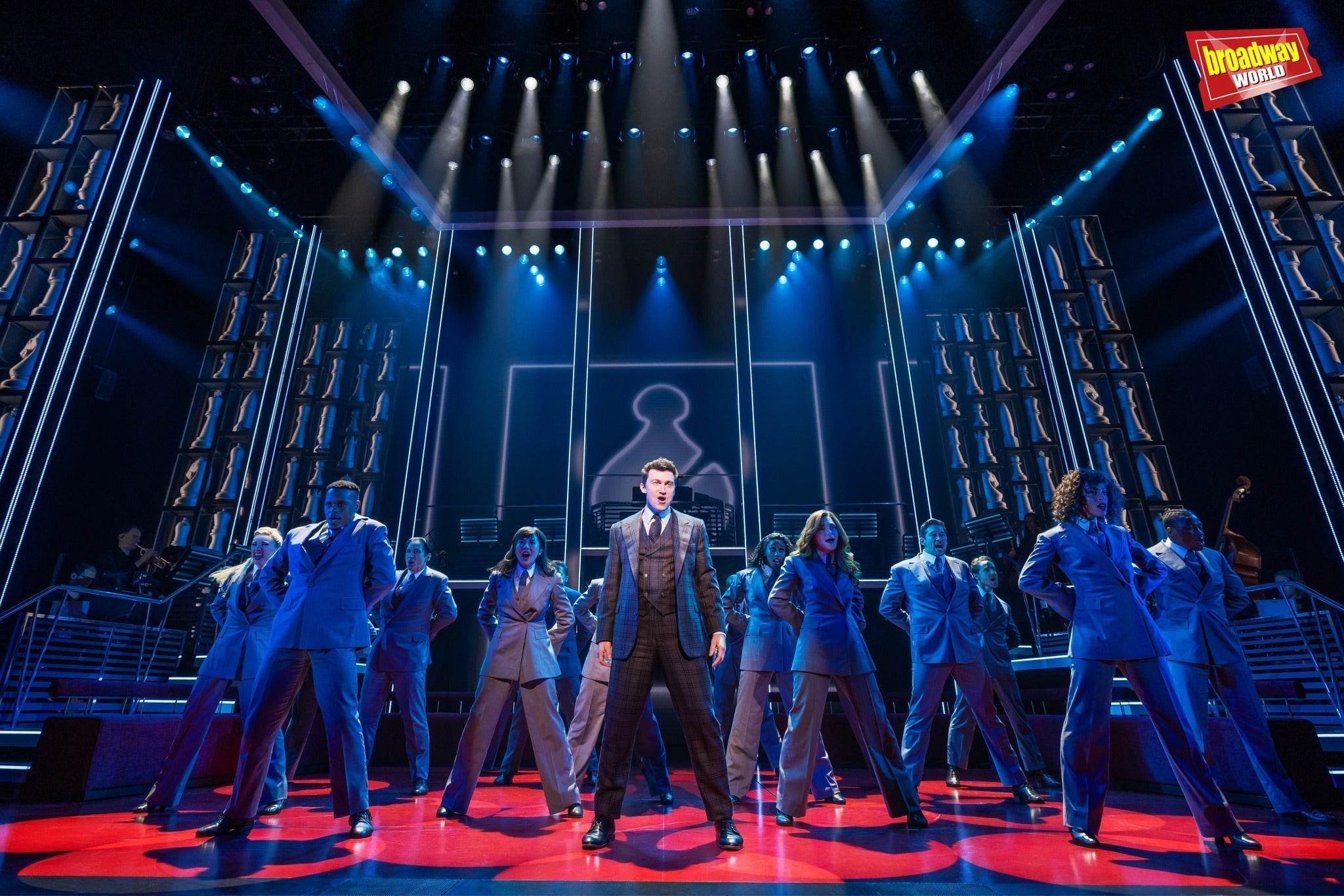 Broadway meets MegaChess! A lively performance with actors in suits on stage under theatrical lighting.