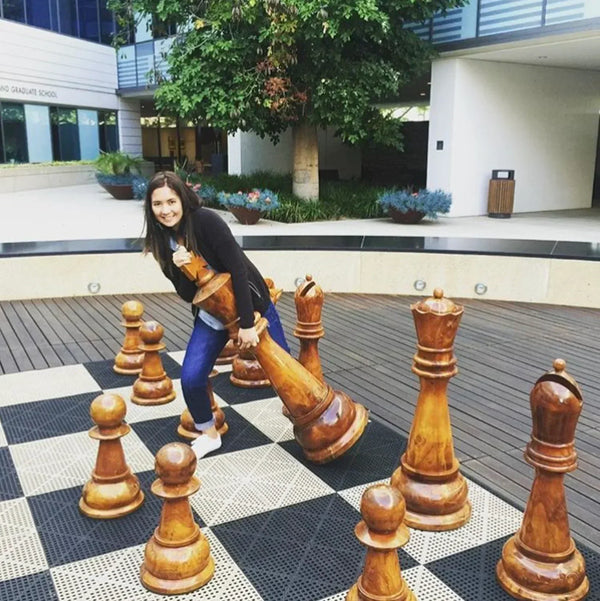 Woman smiling while lifting a giant chess queen on an outdoor board, posing next to large wooden pieces showing why is chess a sport with physical engagement.