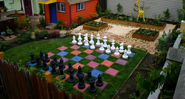 Backyard game area ideas giant chess set on colored grass board in backyard garden next to raised planters and playhouse.