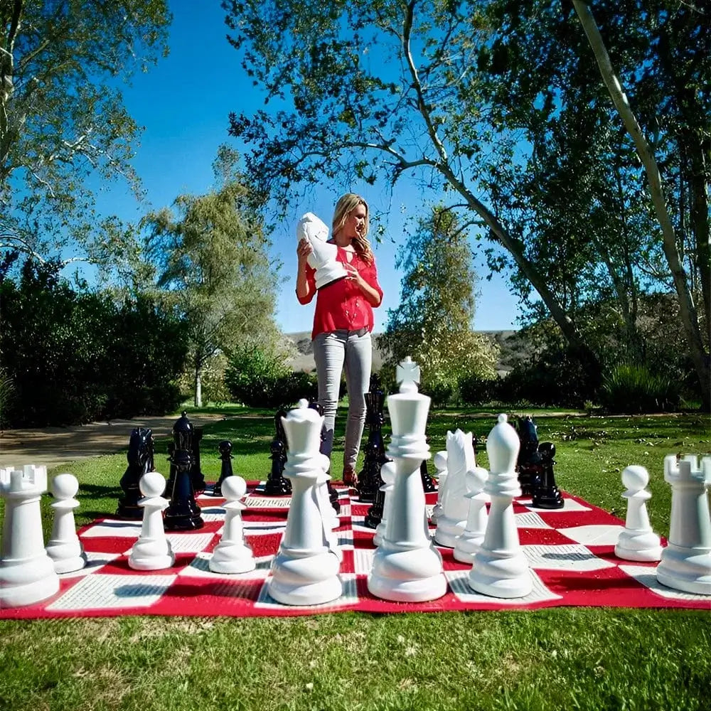 woman-standing-on-red-and-white-giant-chess-board-holding-white-piece-in-grassy-park-with-trees