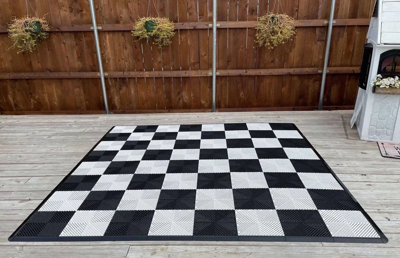 Black and white checkered chessboard laid on wood deck showing a larger version compared to a standard chess board size in an outdoor setting.