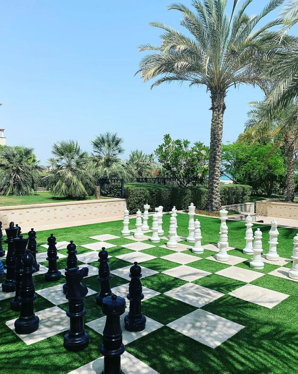 Black and white giant chess set on outdoor checkered board surrounded by palm trees, greenery, and blue sky showcasing a luxury hotel with giant chess board in resort setting.