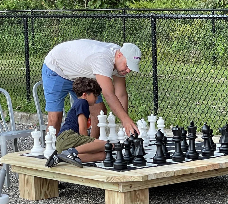 Father and son playing on raised giant chess board outdoors while discussing moves showing how to get better at chess through hands-on guidance.