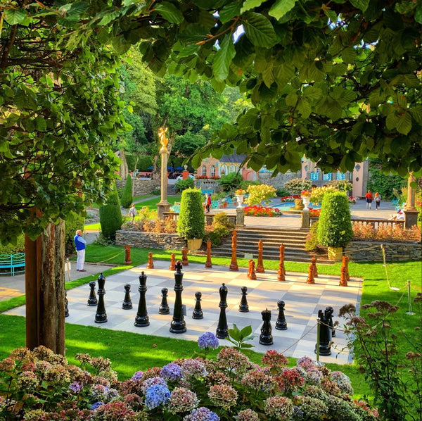 Giant outdoor chessboard with black and brown pieces in beautiful garden surrounded by flowers trees and decorative architecture on a sunny day.