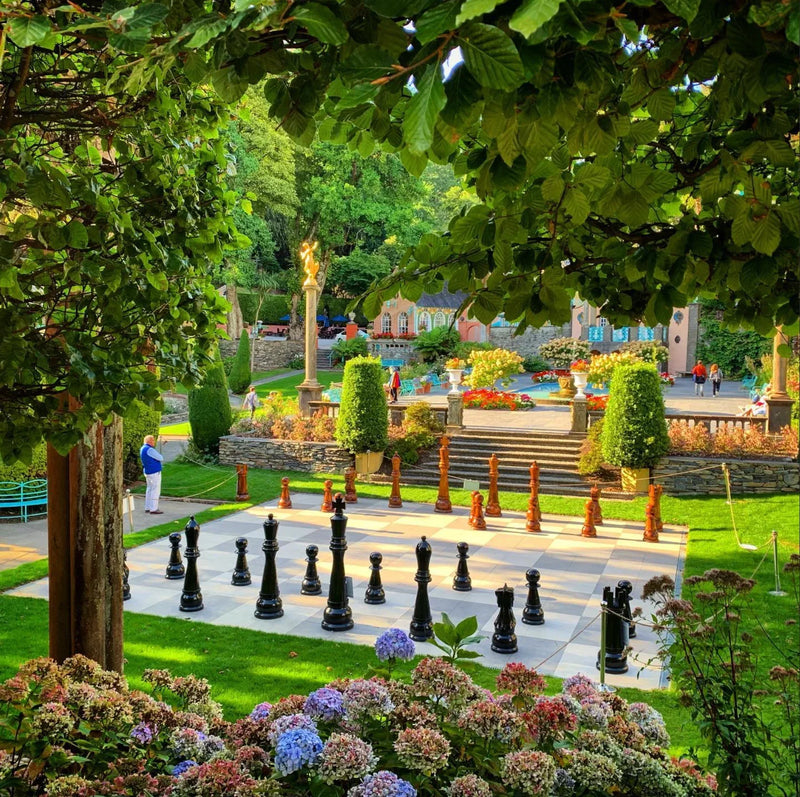Giant outdoor chessboard with black and brown pieces in beautiful garden surrounded by flowers trees and decorative architecture on a sunny day.
