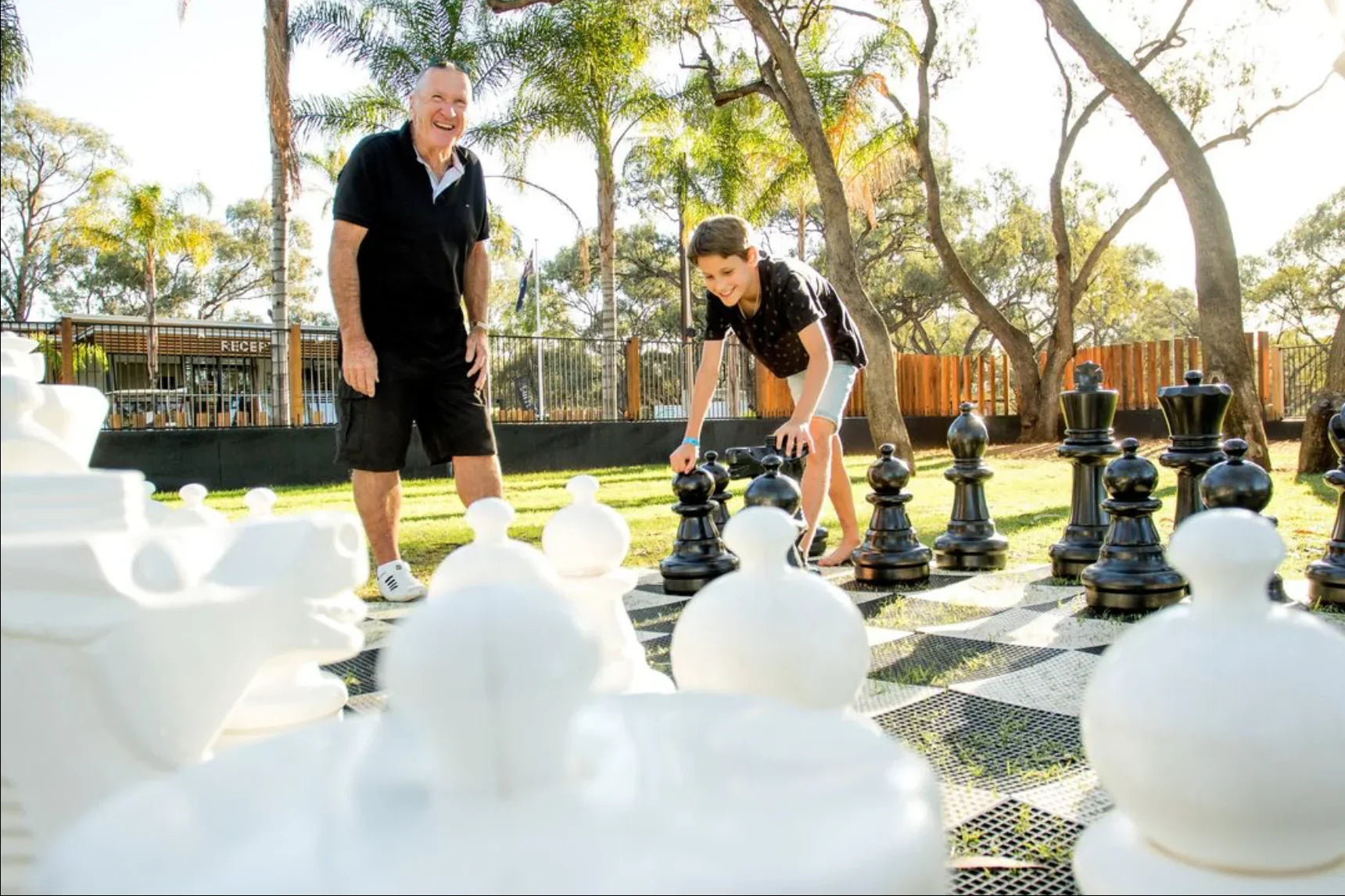 Grandfather and grandson playing giant chess on lawn under sunlight, smiling and engaged in outdoor family party games in a park-like setting.