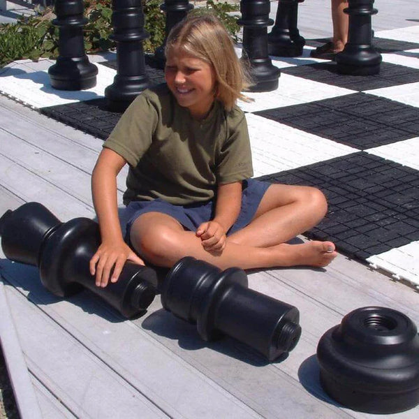 How to make a giant chess set - Child sitting on deck smiling while assembling large black giant chess piece parts next to outdoor chessboard.