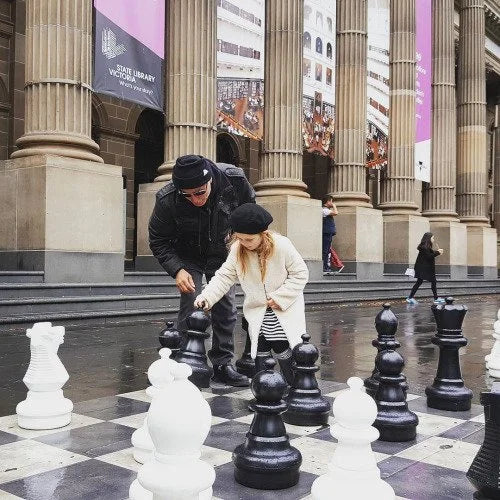 Is chess hard to learn - adult and child playing giant chess outside state library with columns and banners in background on large checkered chessboard.
