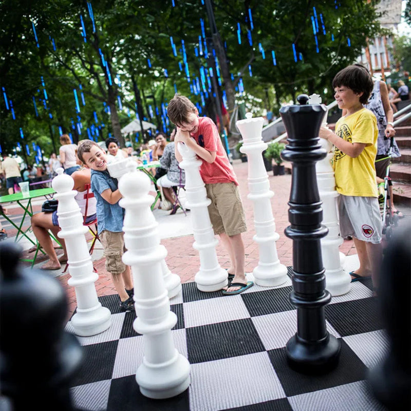 kids-laughing-and-playing-with-giant-chess-pieces-on-an-outdoor-board-in-a-park-while-people-sit-in-the-background-showing-gift-ideas-for-chess-lovers
