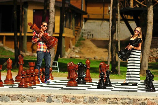 Man and woman holding large chess pieces on outdoor giant chessboard surrounded by trees and buildings in sunny weather.