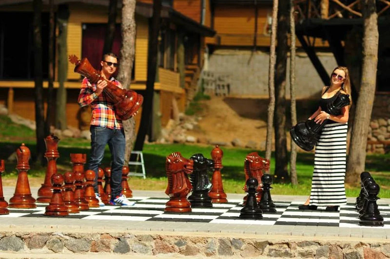 Man and woman holding large chess pieces on outdoor giant chessboard surrounded by trees and buildings in sunny weather.