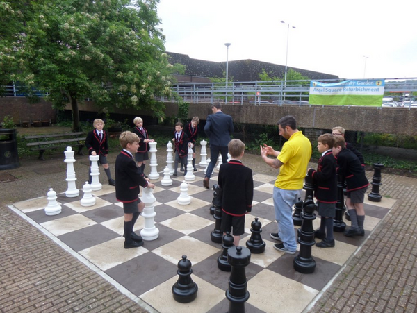 What does chess teach you children and adults playing giant chess outdoors on checkered board at school yard in uniforms.