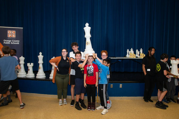 why-are-some-kids-good-at-chess-group-of-kids-and-adults-smiling-while-holding-a-giant-white-chess-piece-on-a-school-stage