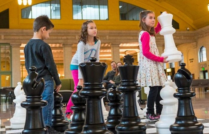 Children playing with giant black and white chess pieces on indoor board demonstrating use of giant chess set for schools in an educational setting.