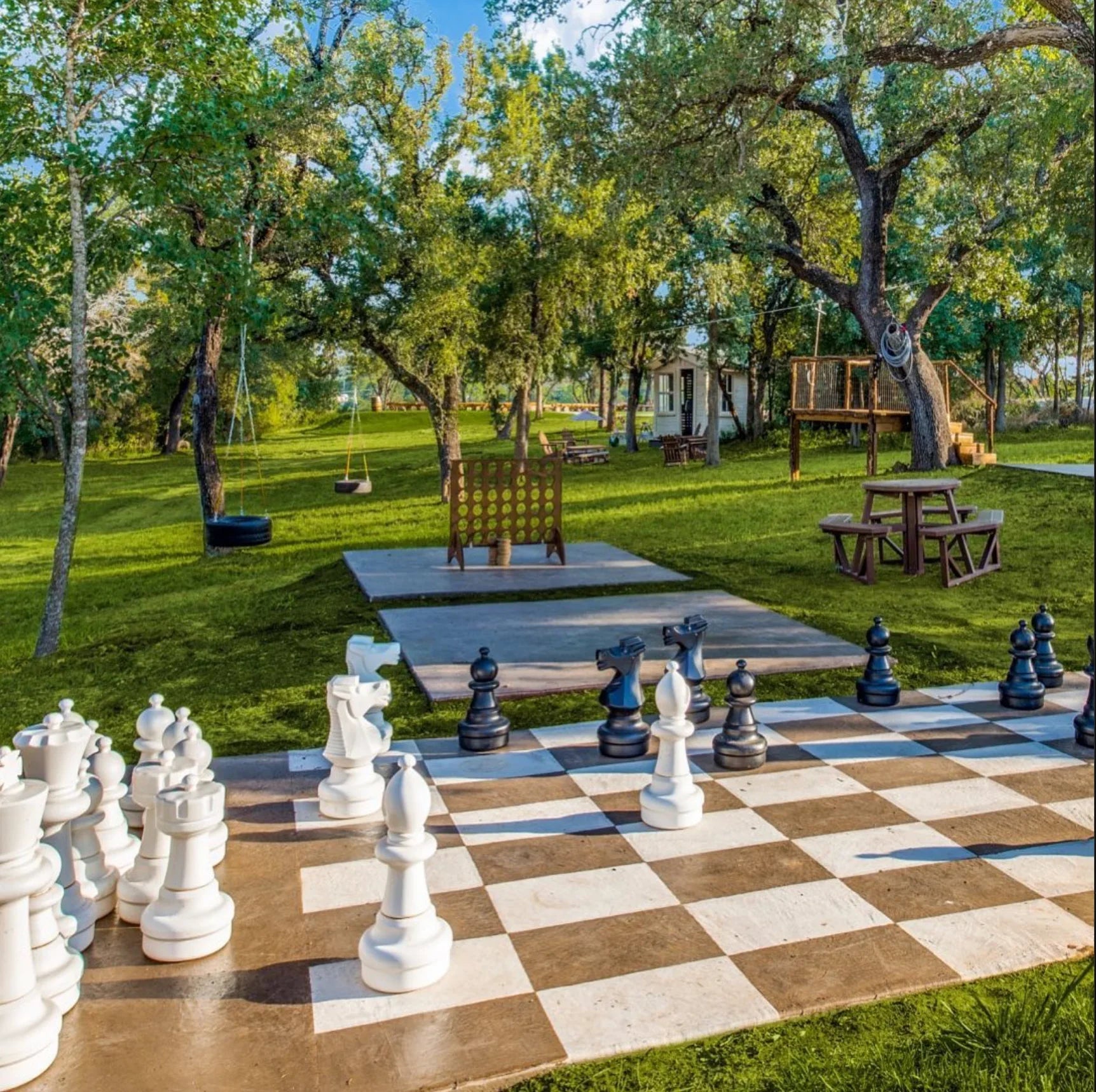 giant-chess-set-and-4-in-a-row-lawngames-arranged-on-concrete-pads-in-a-green-park-with-picnic-tables-tire-swings-and-trees
