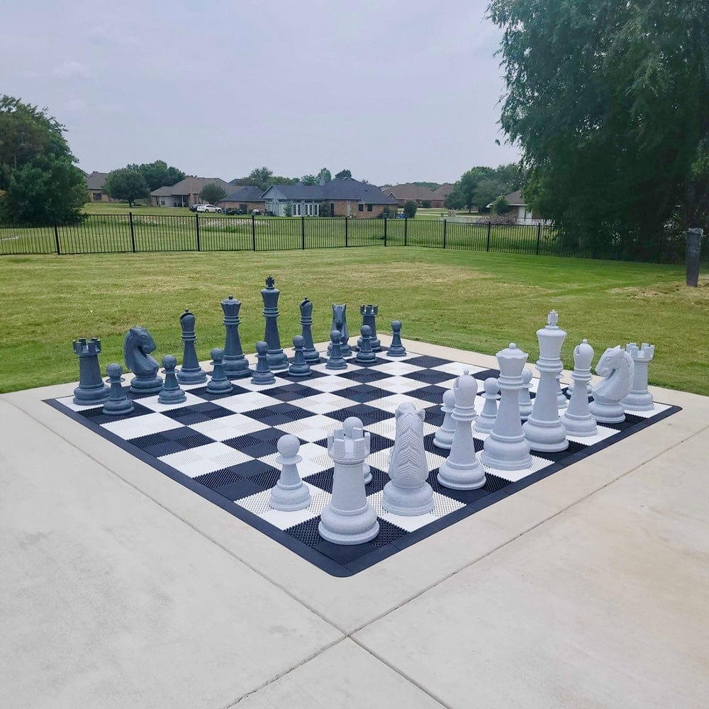 giant-outdoor-chess-set-stone-gray-pieces-on-black-and-white-board-placed-on-concrete-surface-with-green-grass-lawns-trees-and-houses-in-background