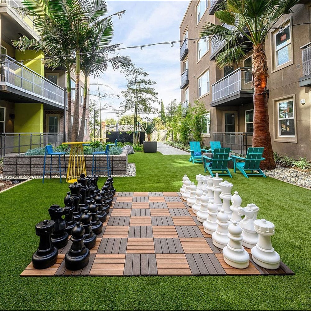 Black and white plastic chess set arranged for gameplay on large outdoor checkered board with planter boxes and tables in background under sunny sky.
