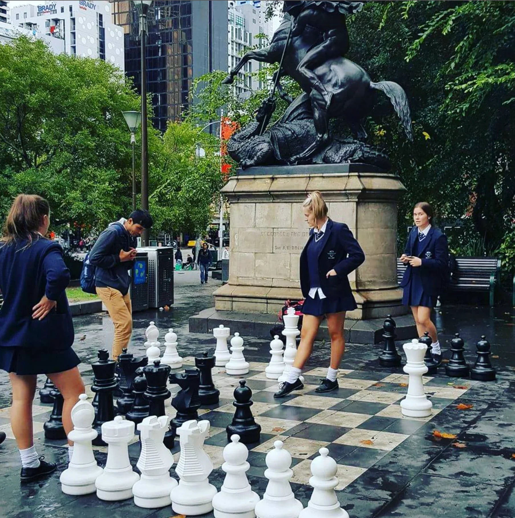 students-in-uniform-playing-oversized-outdoor-chess-set-in-city-square-next-to-large-horse-statue-surrounded-by-trees-and-tall-buildings