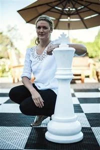 Woman kneeling on outdoor black and white checkered board resting hand on giant white king chess piece with umbrella and seating area in background.